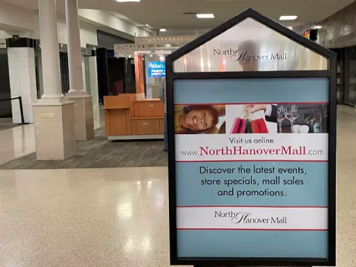 A sign encourages people to visit www.NorthHanoverMall.com to learn about current events, store deals, and shower installation offers from Mid State Baths. The setting shows a quiet shopping mall. Alt text: Sign promotes Mid State Baths shower offers and events at North Hanover Mall website.