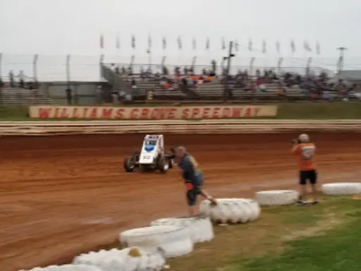 A race car moves quickly on the dirt track at Williams Grove Speedway as fans watch and photographers take pictures near Mid State Baths banners. Alt text: Race car passes Mid State Baths banners on dirt track with fans and photographers watching.